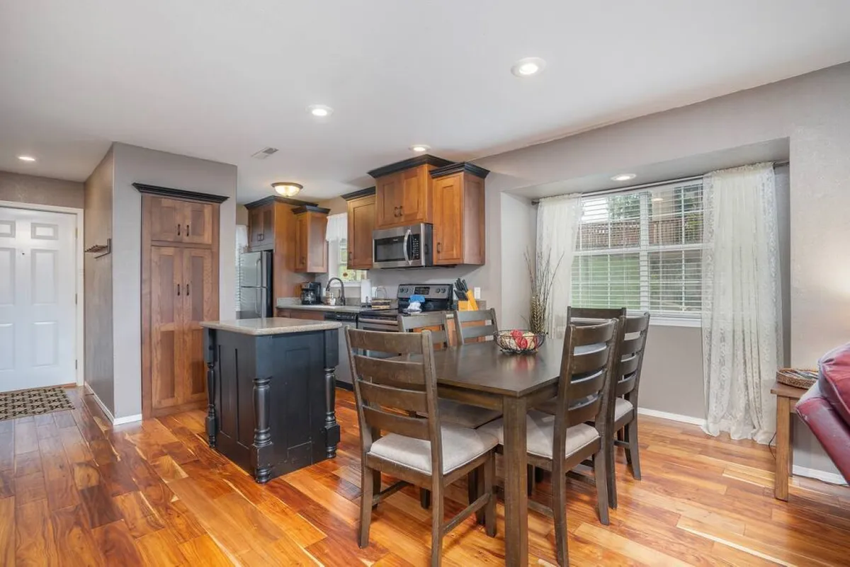 A cozy and welcoming dining area seamlessly connected to the kitchen, featuring a wooden dining table, a kitchen island, and a window with curtains. The setup invites family meals, entertaining, and intimate conversations, reflecting a harmonious blend of cooking and dining spaces.