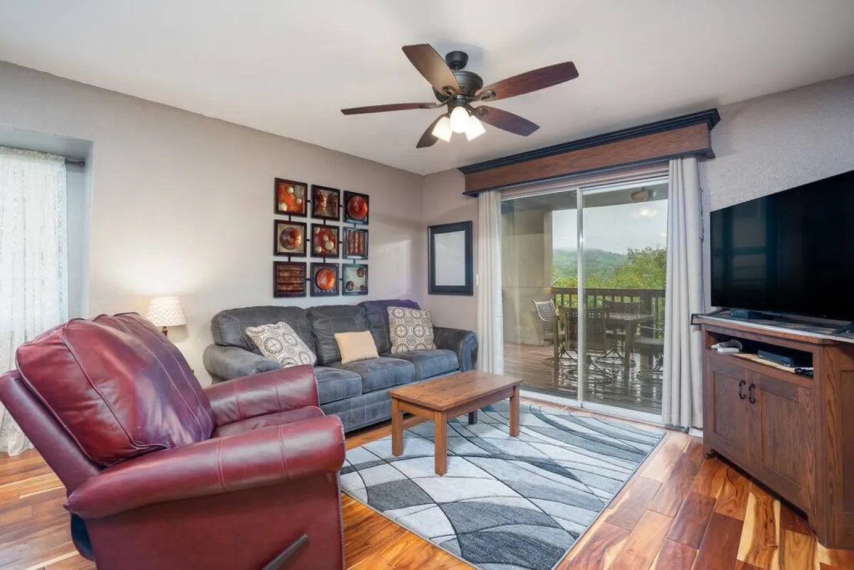 A cozy living room featuring a comfortable couch, recliner, and coffee table, with artwork adorning the wall. Large sliding doors open to a balcony, allowing natural light to flood the space and offering potential views outside.