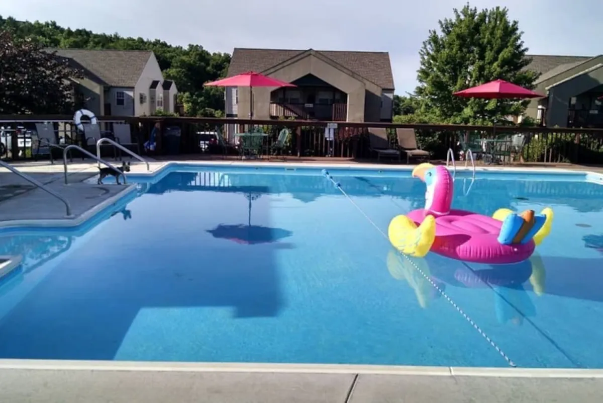 A large outdoor swimming pool is surrounded by bright red umbrellas and lounge chairs, with playful touches like a flamingo float. The pool is bordered by attractive community buildings, creating a lively and inviting environment ideal for socializing or relaxing under the sun.