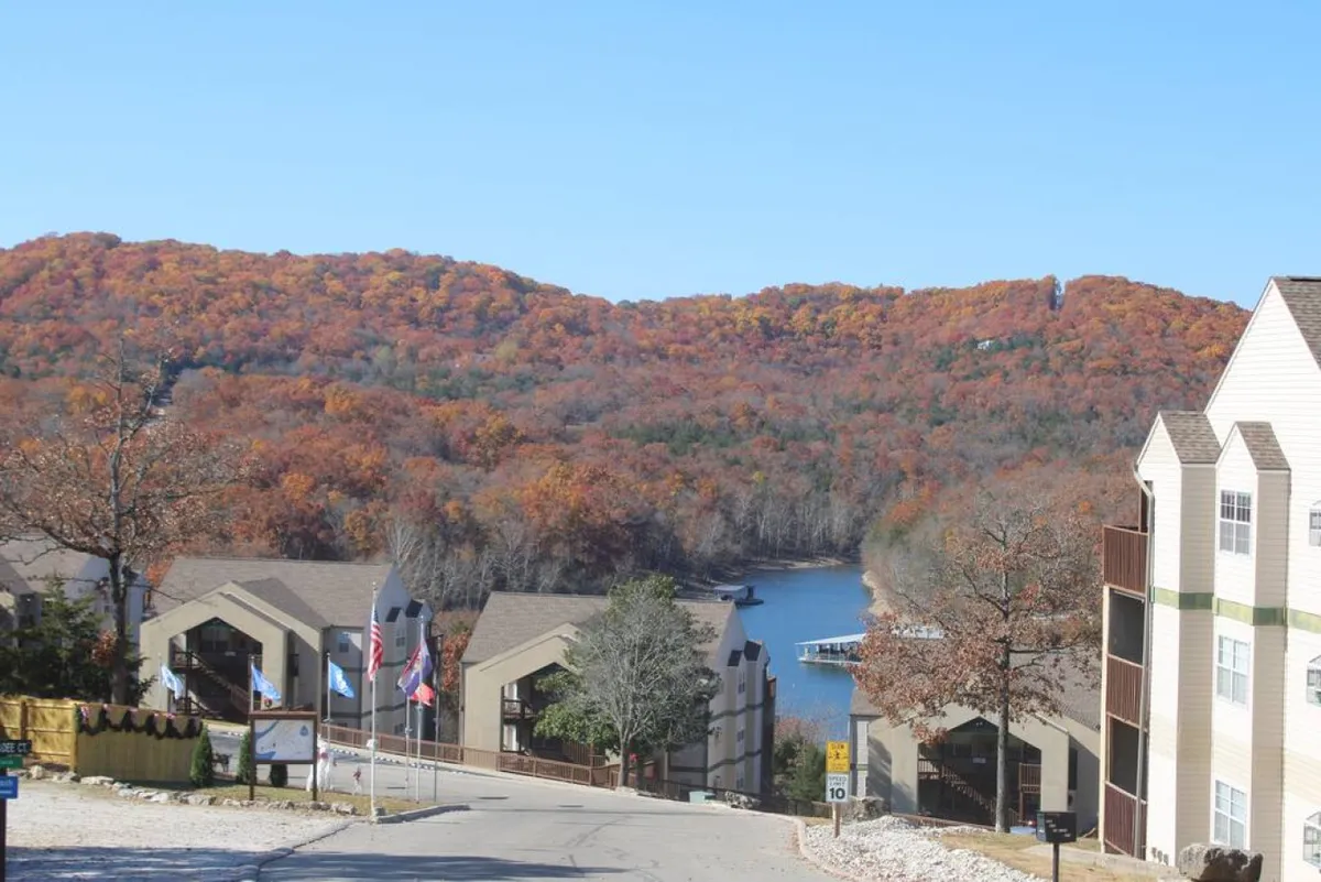 A broad lakeside vista with rich autumn leaves in the trees, a clear view of the lake, and glimpses of modern resort buildings. The scene is alive with color, showcasing both the natural environment and the lakeside amenities available to visitors.