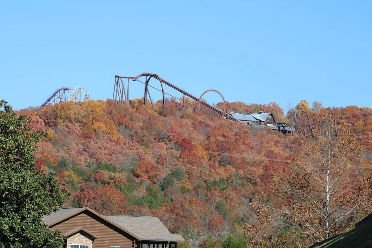 The exterior scene showcases charming houses nestled among vibrant autumn foliage, with the thrill of a nearby roller coaster visible in the distance. The image captures the essence of Branson's family-friendly attractions and stunning seasonal scenery, highlighting the property's close proximity to Silver Dollar City and natural beauty.