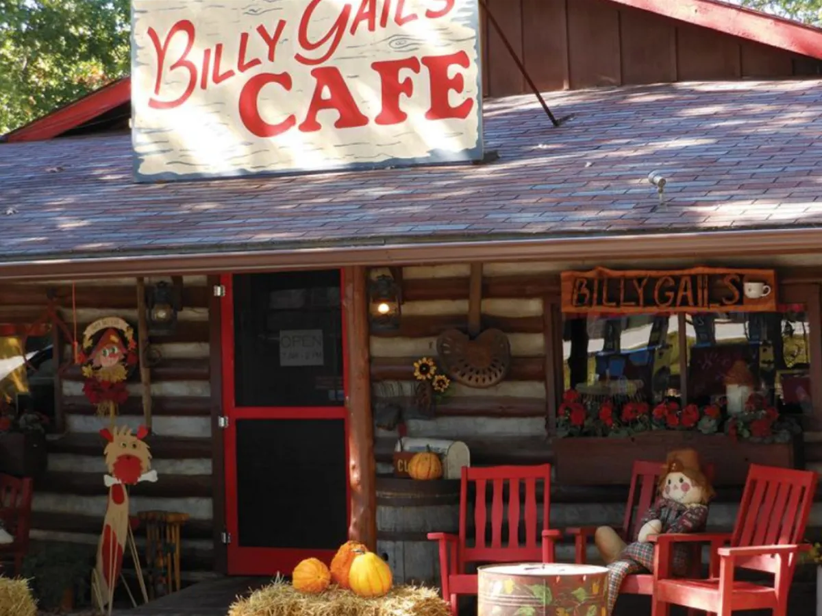 The image showcases a cheerful café exterior with a welcoming sign, red chairs, pumpkins, and a festive scarecrow decoration. The setting suggests a lively social hub for dining, socializing, and experiencing local charm, accented by seasonal touches.