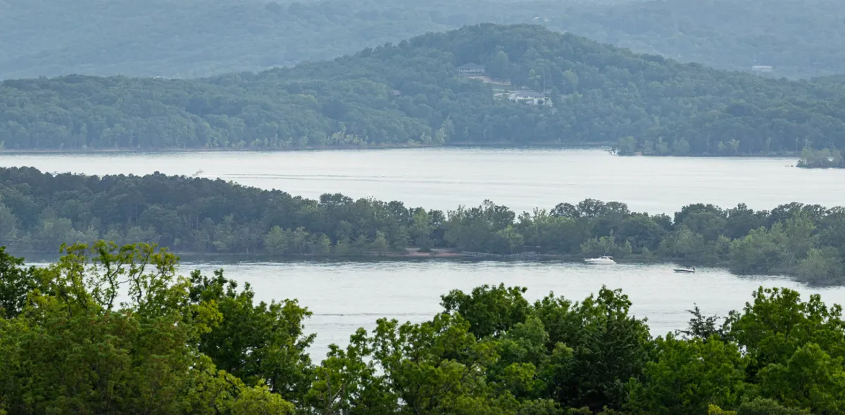 A peaceful lakeside scene featuring the glistening surface of Table Rock Lake, tree-lined shores, and distant mountains. The calm water invites activities like boating, fishing, or simply relaxing by the shoreline.