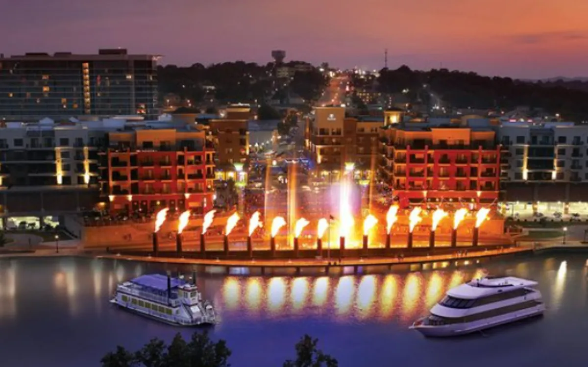 A dynamic evening waterfront scene with illuminated water features, boats, colorful buildings, and a lively atmosphere. People are dining along the waterfront, enjoying live entertainment, and the area is vibrant under the evening sky.